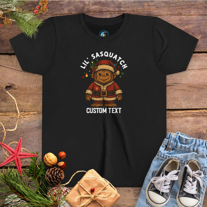 Black t-shirt with Christmas-themed graphic and 'Lil Sasquatch' text on a wooden surface with festive decorations.