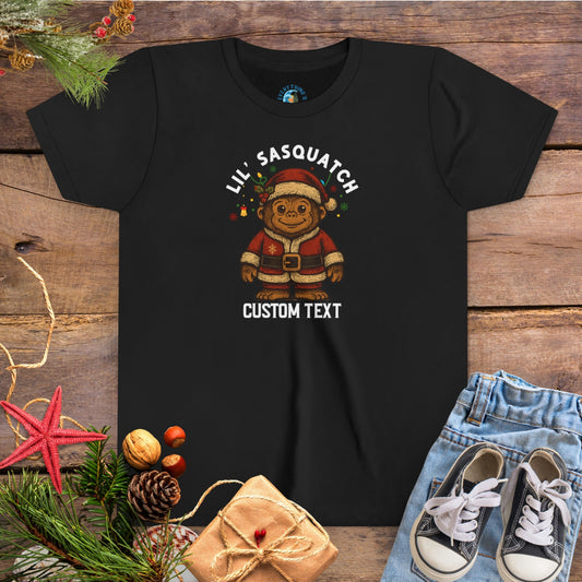 Black t-shirt with Christmas-themed graphic and 'Lil Sasquatch' text on a wooden surface with festive decorations.