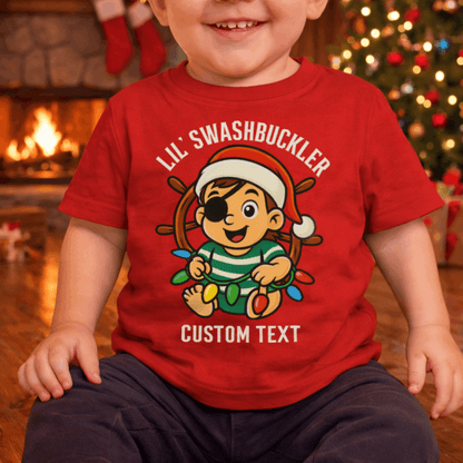 Child wearing a red t-shirt with a pirate-themed design and 'Lil' Swashbuckler' text, sitting in front of a Christmas tree and fireplace.