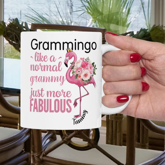 Ceramic coffee mug featuring a flamingo adorned with flowers and the text “Grammingo like a normal Grammy just more fabulous,” available with white, pink, or black handle options.