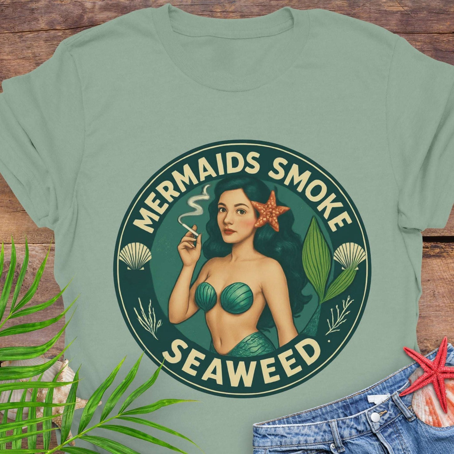 Green t-shirt with mermaid graphic and text 'Mermaids Smoke Seaweed' on a wooden surface.