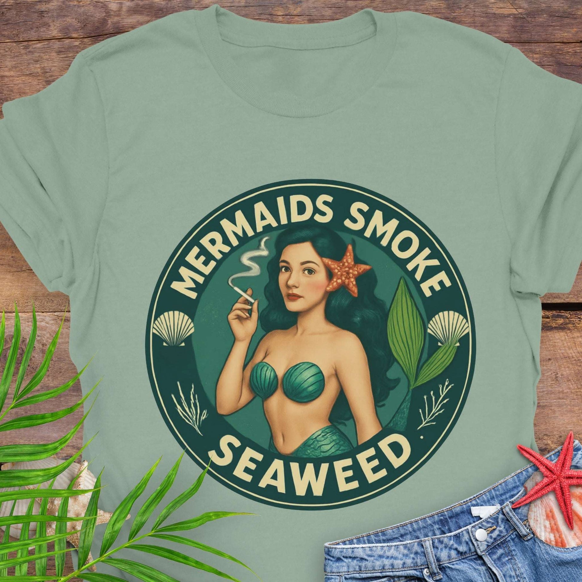 Green t-shirt with mermaid graphic and text 'Mermaids Smoke Seaweed' on a wooden surface.