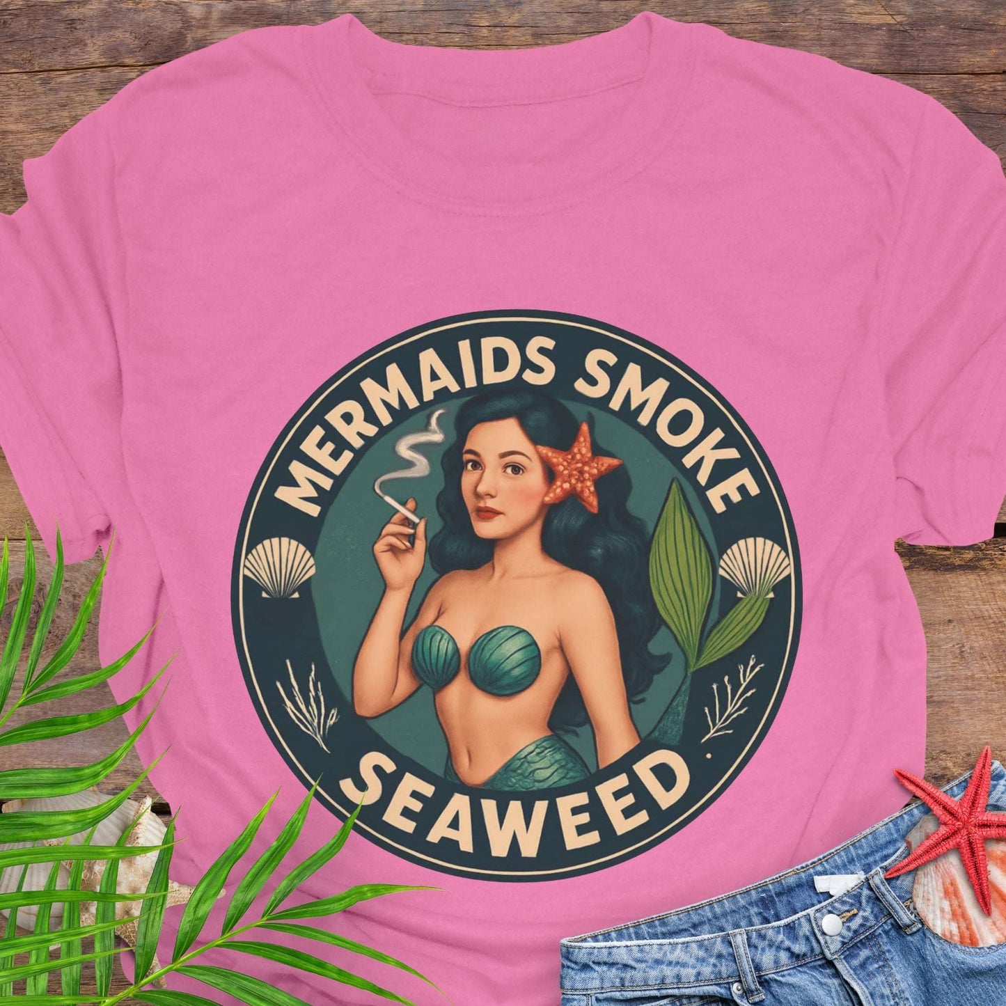 Pink t-shirt with mermaid graphic and 'Mermaids Smoke Seaweed' text on a wooden surface.