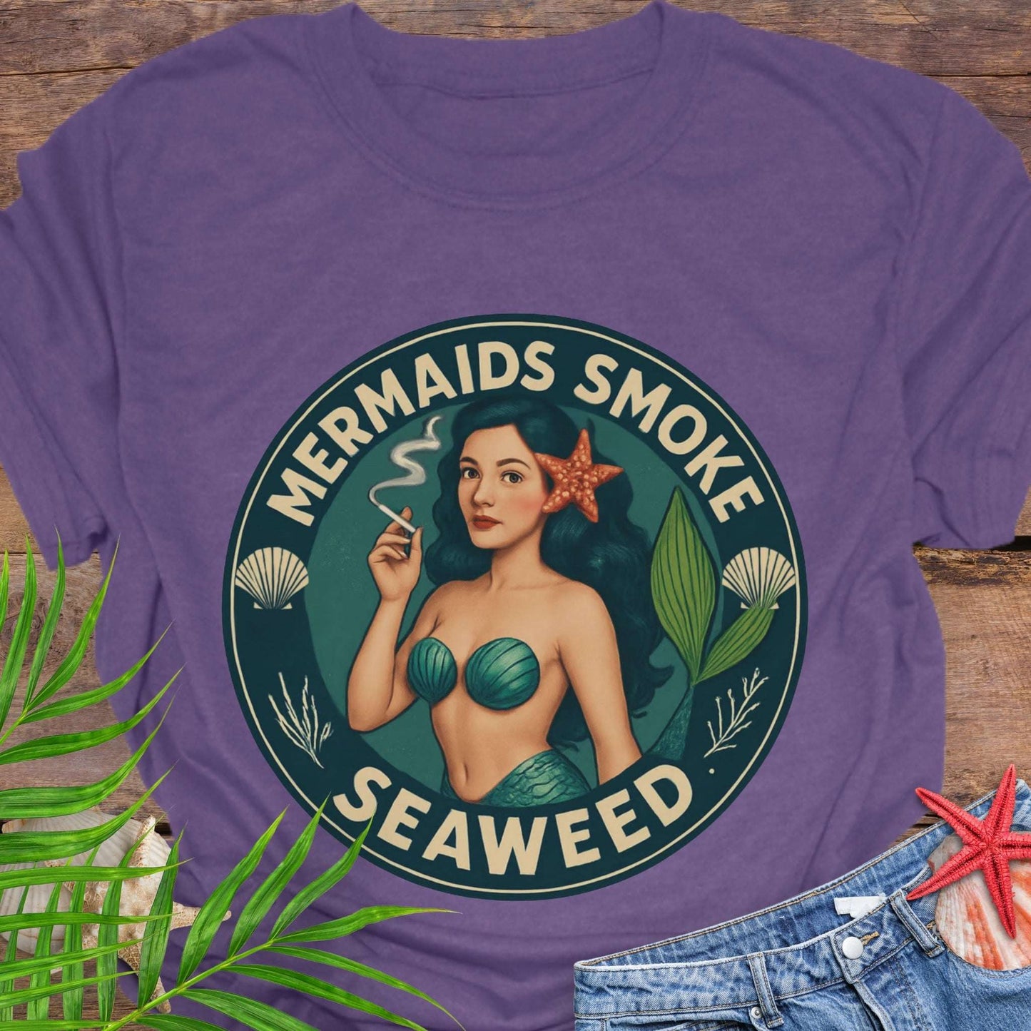 Purple t-shirt with mermaid graphic and 'Mermaids Smoke Seaweed' text on a wooden surface.