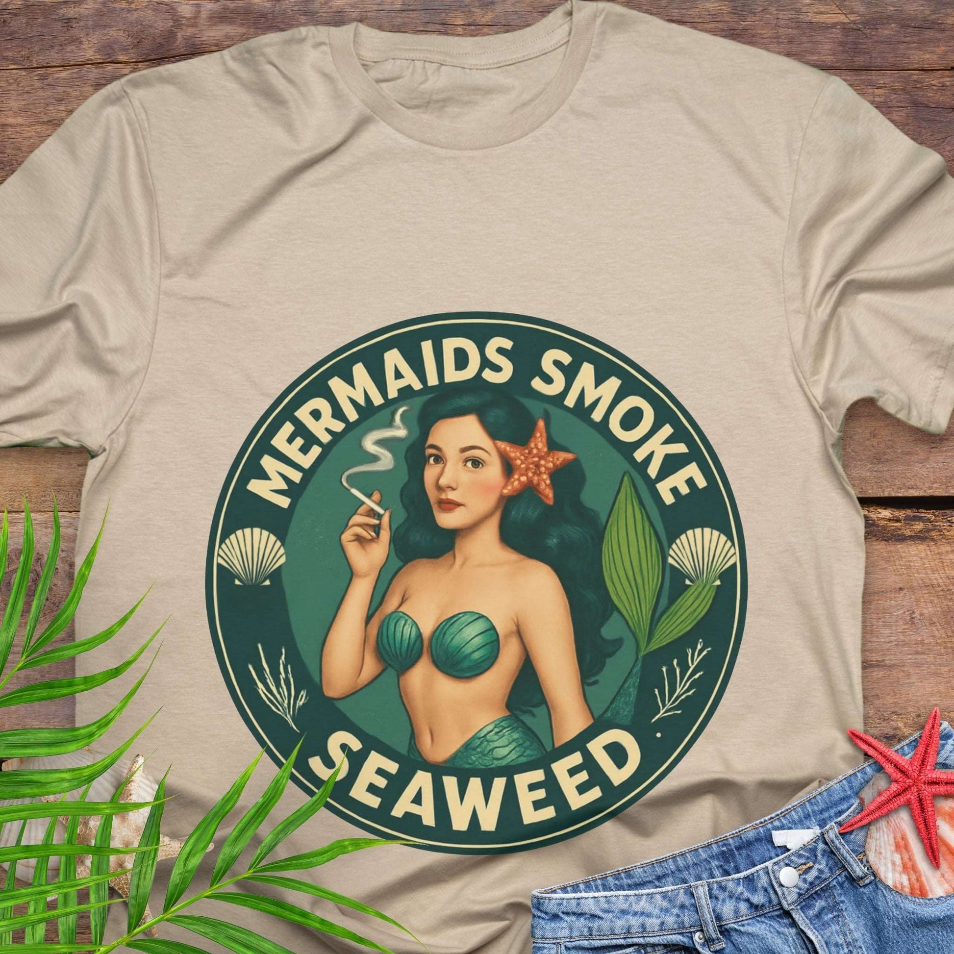 Beige t-shirt with mermaid graphic and text 'Mermaids Smoke Seaweed' on a wooden surface.