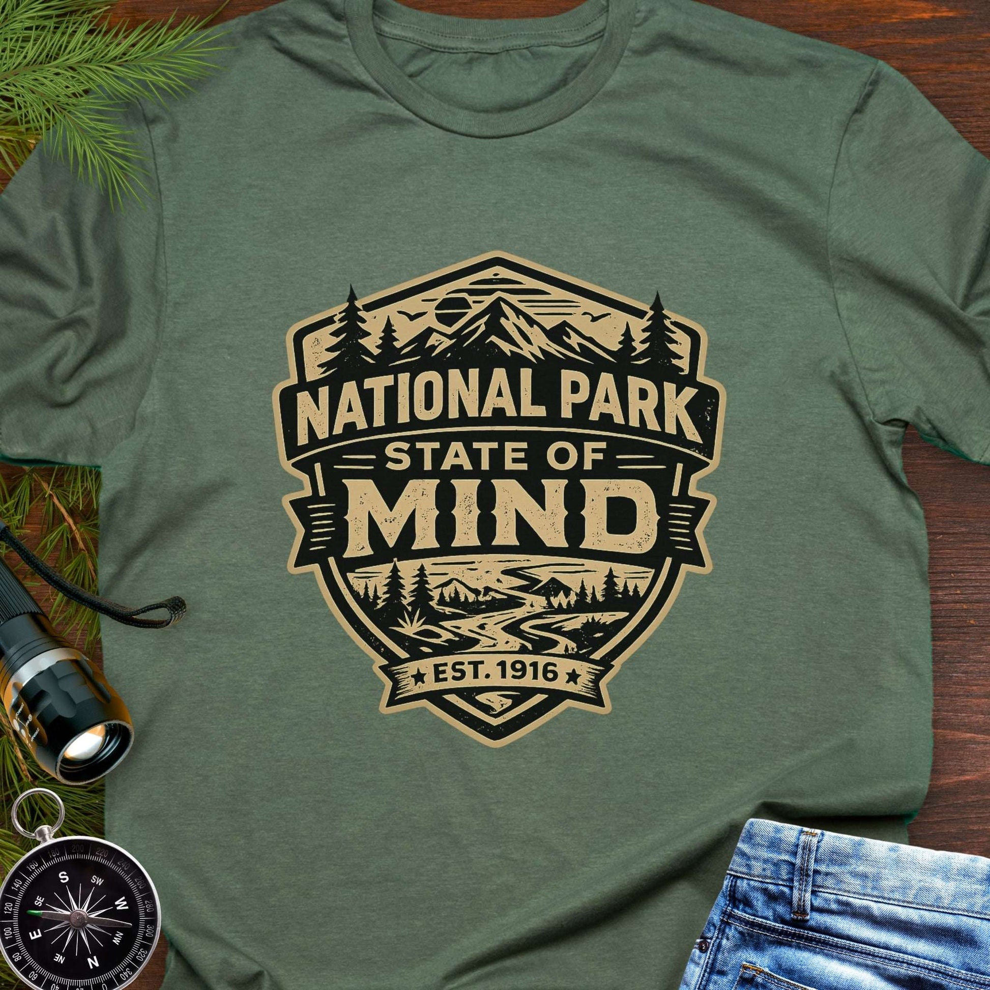 Military Green outdoor t-shirt flat lay with vintage badge graphic reading “National Park State of Mind” and “Est. 1916,” featuring mountains, trees, and a winding river.
