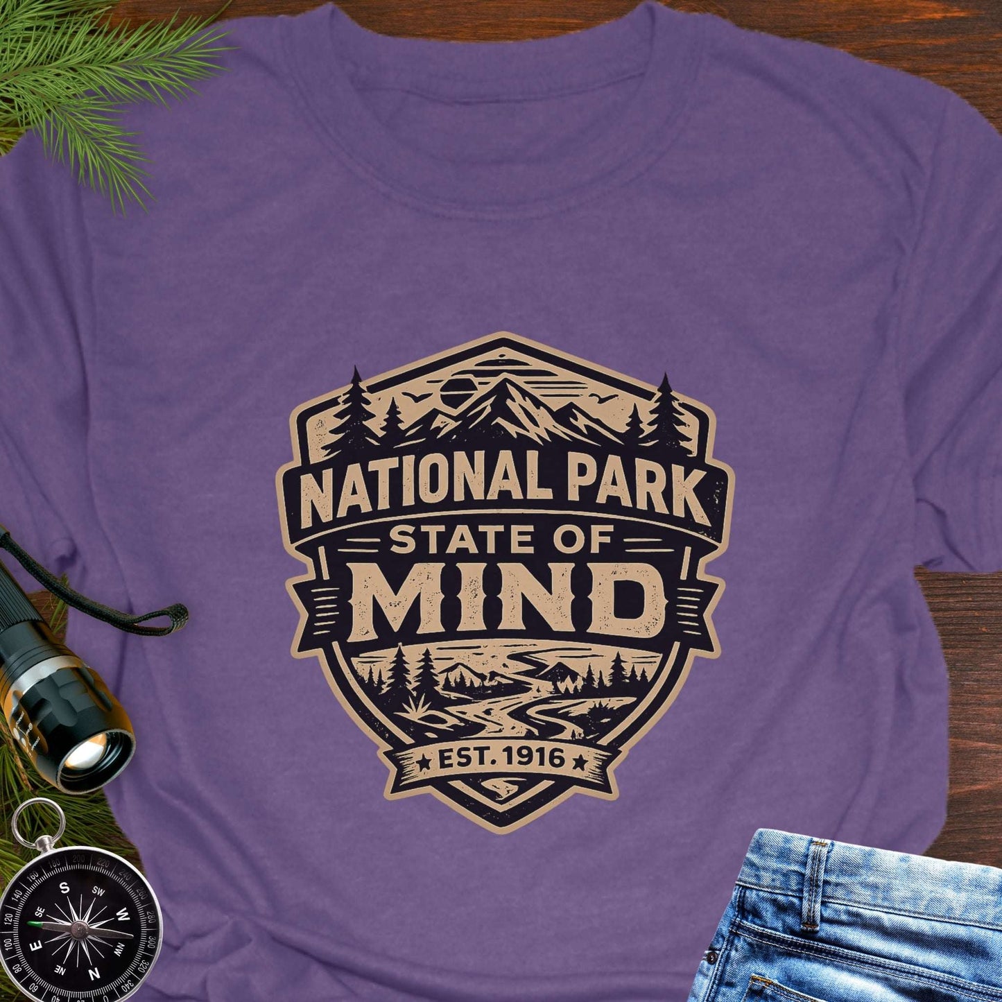 Purple outdoor t-shirt flat lay with vintage badge graphic reading “National Park State of Mind” and “Est. 1916,” featuring mountains, trees, and a winding river.