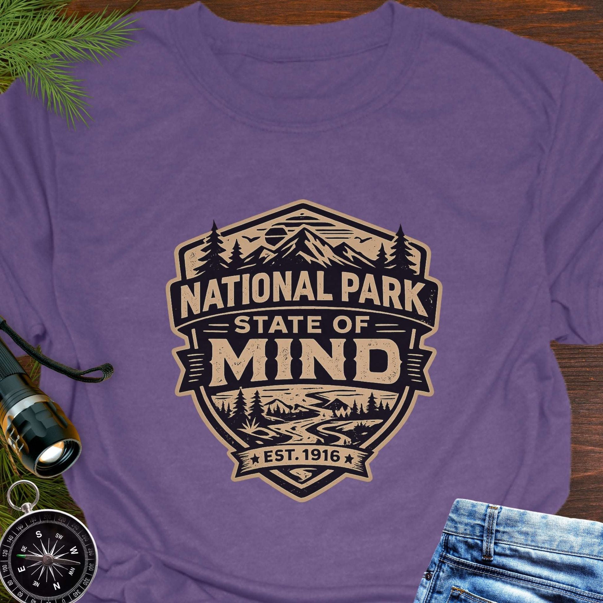 Purple outdoor t-shirt flat lay with vintage badge graphic reading “National Park State of Mind” and “Est. 1916,” featuring mountains, trees, and a winding river.