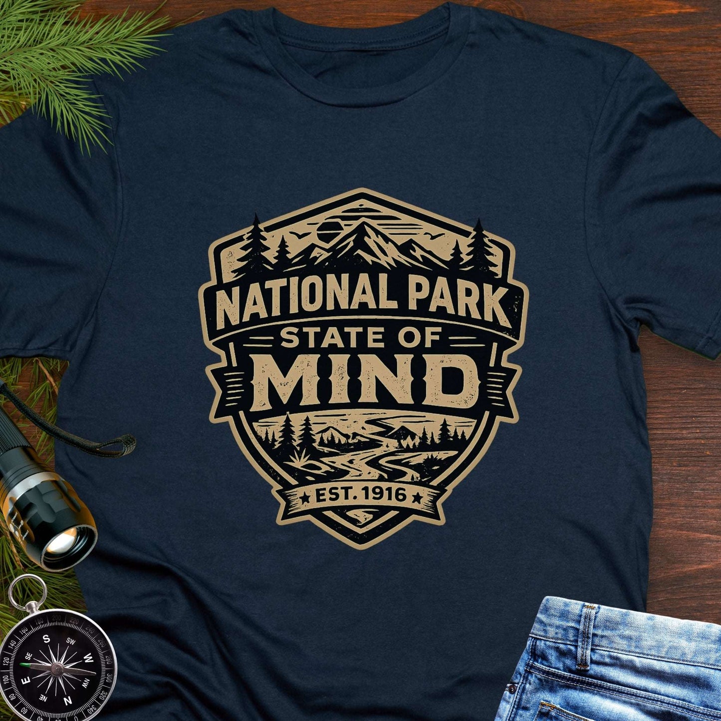 Navy blue outdoor t-shirt flat lay with vintage badge graphic reading “National Park State of Mind” and “Est. 1916,” featuring mountains, trees, and a winding river.