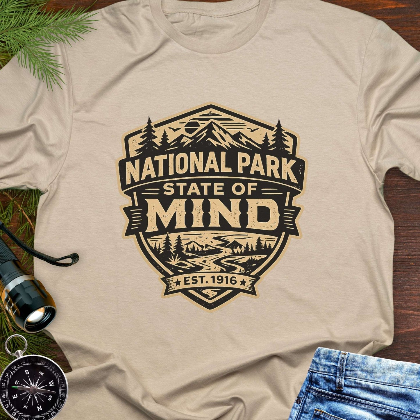 Beige outdoor t-shirt flat lay with vintage badge graphic reading “National Park State of Mind” and “Est. 1916,” featuring mountains, trees, and a winding river.