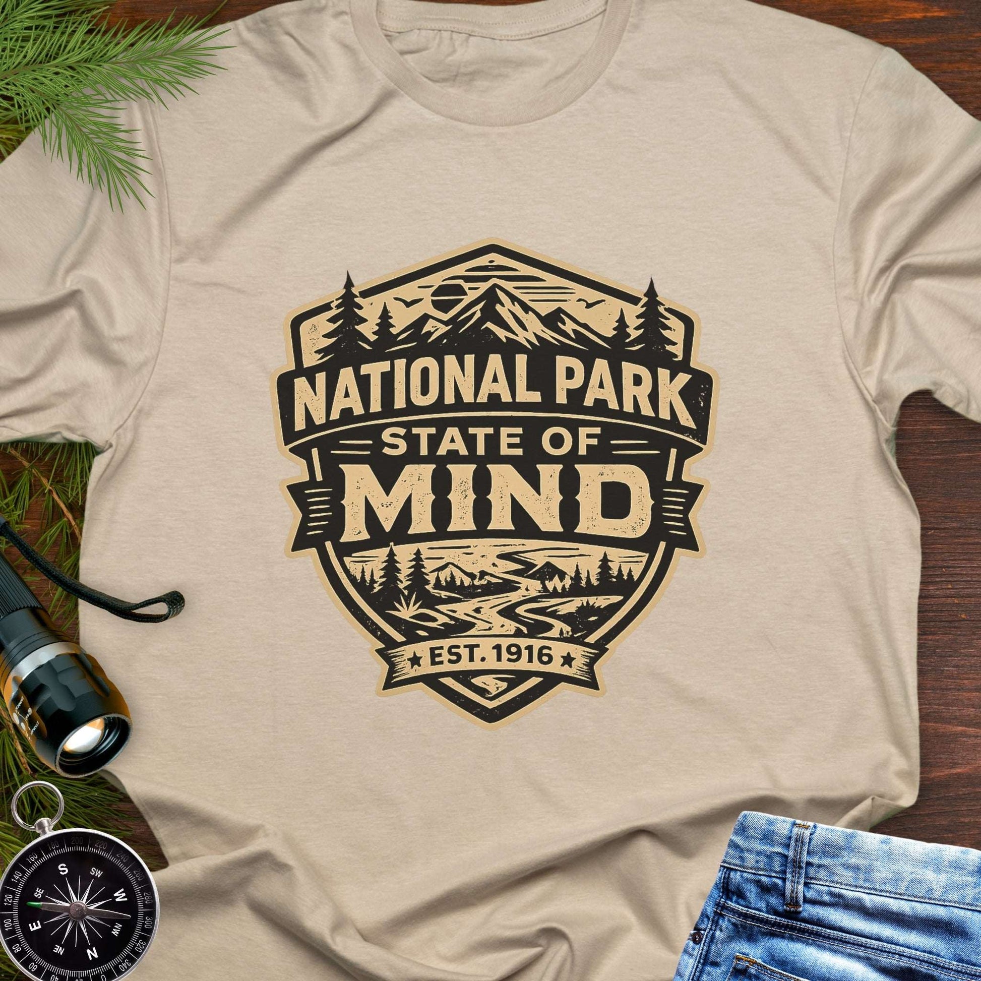 Beige outdoor t-shirt flat lay with vintage badge graphic reading “National Park State of Mind” and “Est. 1916,” featuring mountains, trees, and a winding river.