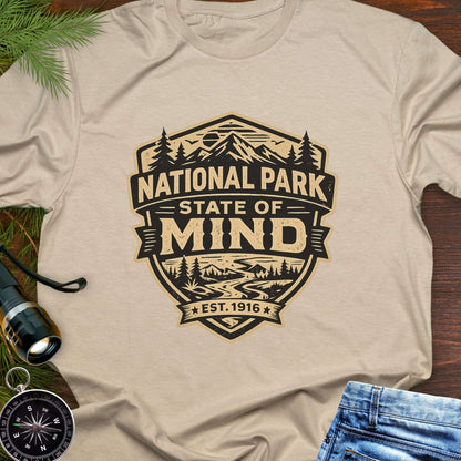 Beige outdoor t-shirt flat lay with vintage badge graphic reading “National Park State of Mind” and “Est. 1916,” featuring mountains, trees, and a winding river.