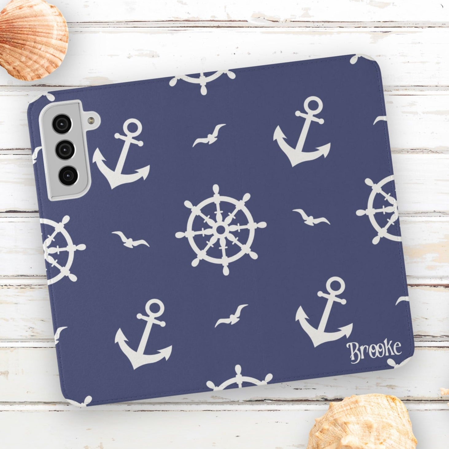 Phone case with nautical design featuring anchors and wheels on a blue background, placed on a wooden surface with shells.