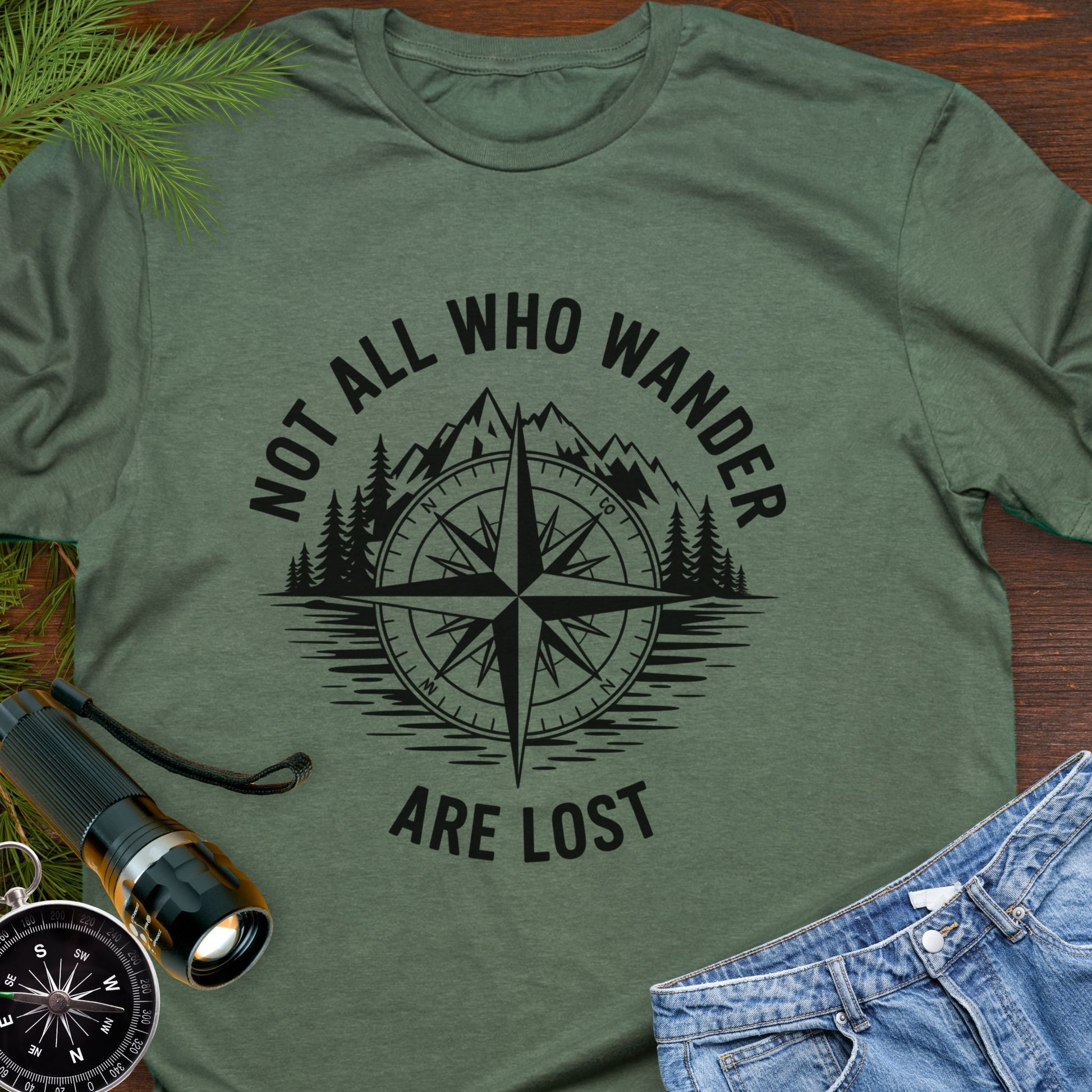 Military Green  T-shirt with a mountain range and lake with a compass overlay that says Not All Who Wander are Lost. 