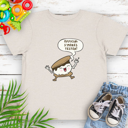 Heather Dust Toddler T-shirt with "Official S'mores Tester" text in speech bubble over a S'more character – perfect for camping and summer fun.