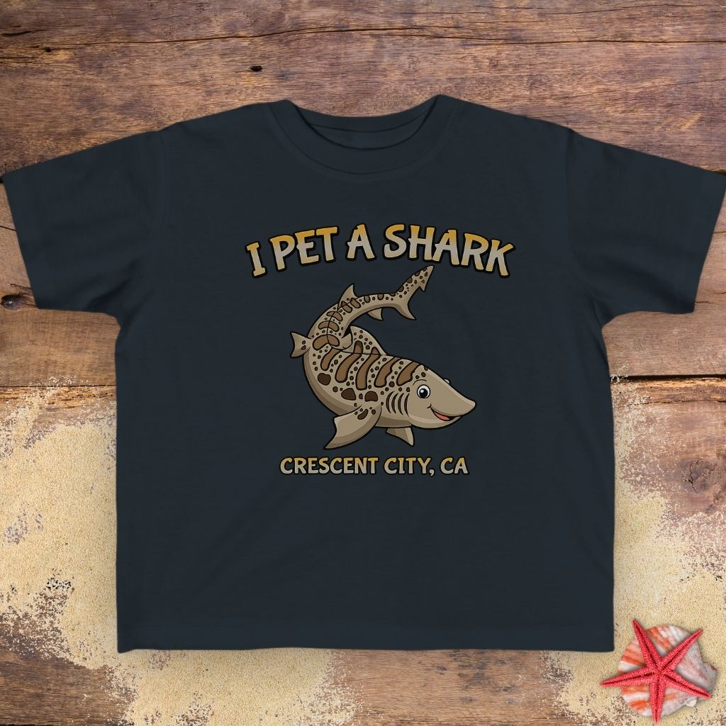 A black short-sleeve t-shirt featuring a graphic of a striped shark with the text 'I PET A SHARK' above it and 'CRESCENT CITY, CA' below it.