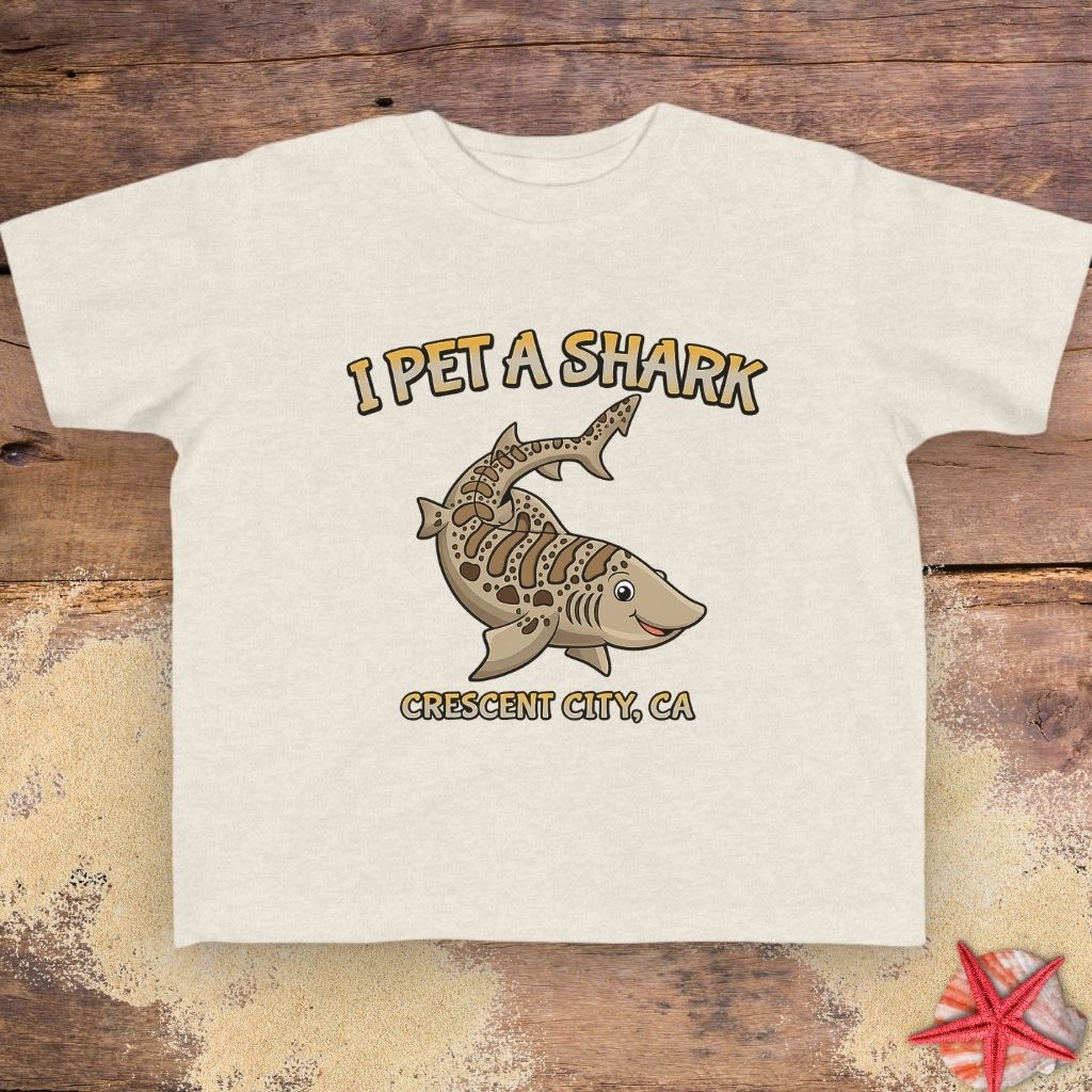 A natural beige short-sleeve t-shirt featuring a graphic of a striped shark with the text 'I PET A SHARK' above it and 'CRESCENT CITY, CA' below it.