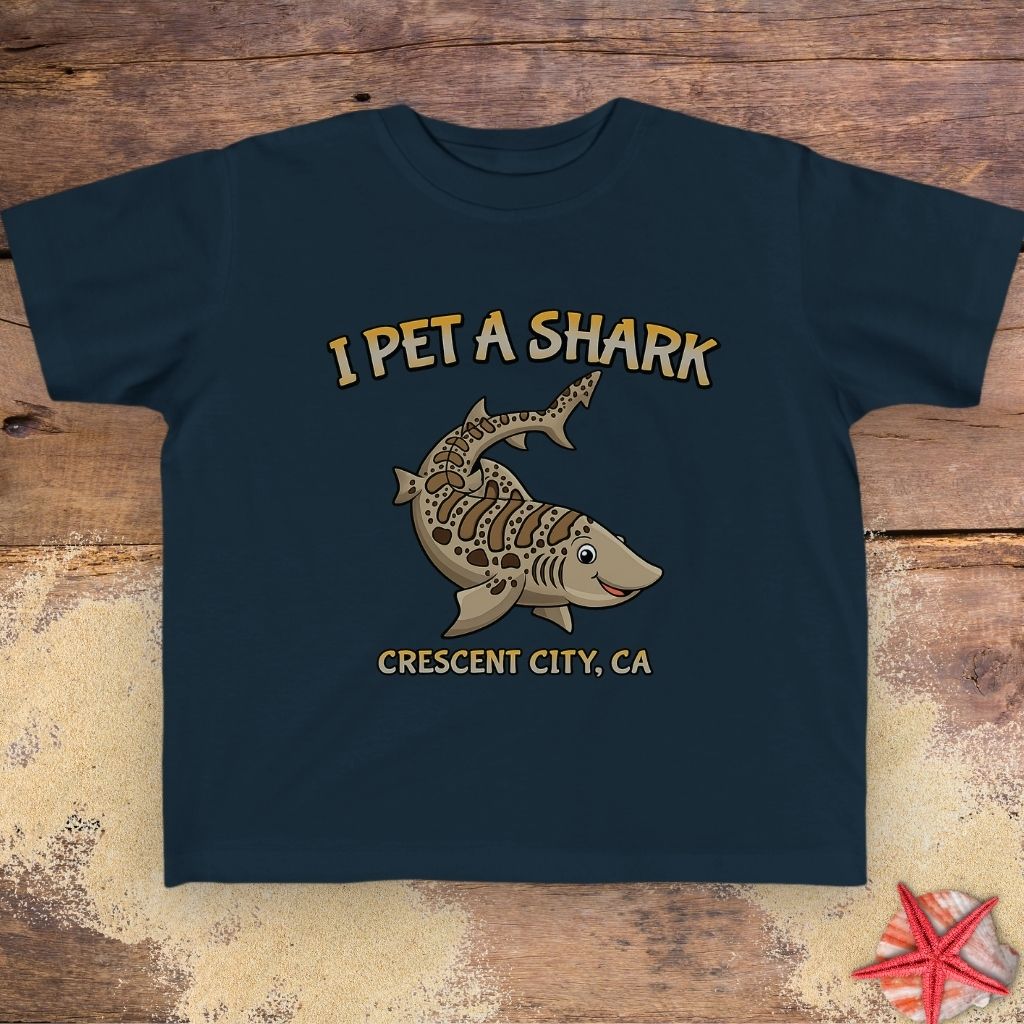A dark navy blue kids' t-shirt featuring a graphic of a striped shark with the text 'I PET A SHARK' above it and 'CRESCENT CITY, CA' below it.