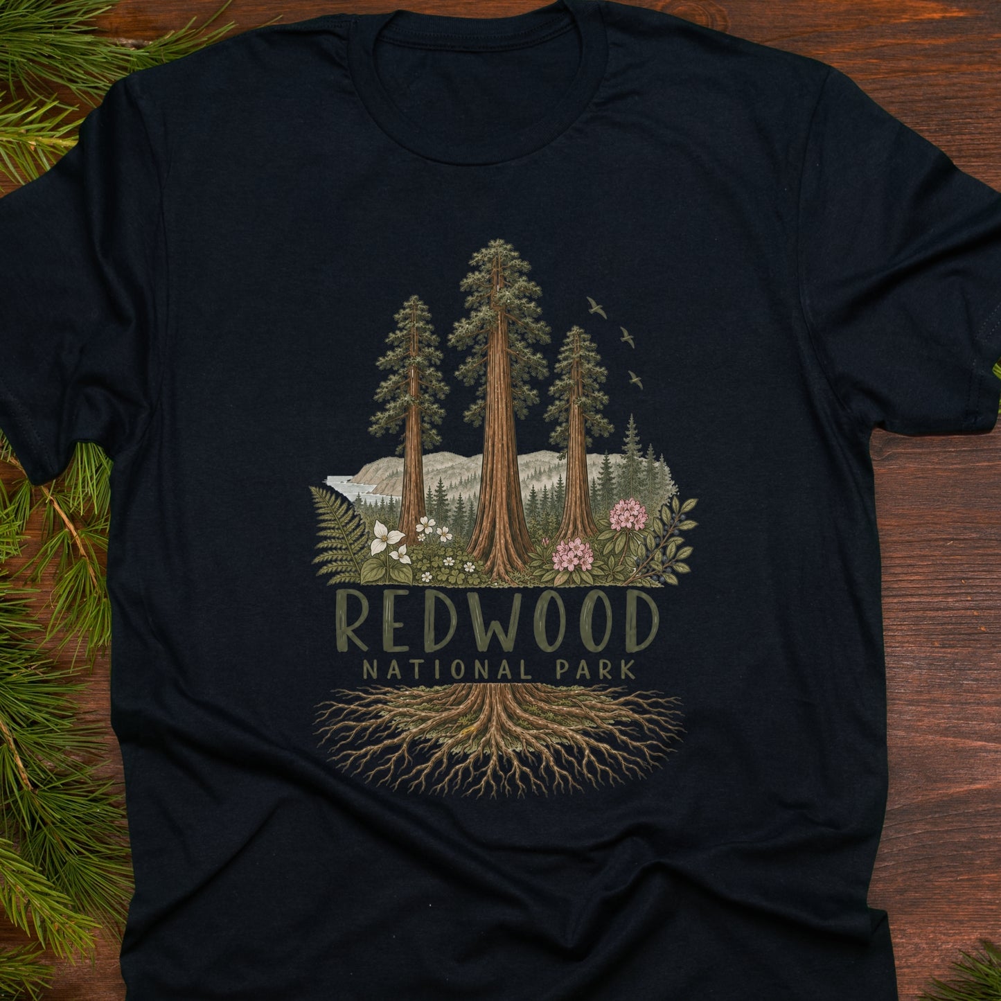 A black short-sleeve t-shirt laid flat on a dark wood surface, decorated with pine branches at the edges. The shirt features a detailed graphic of three tall redwood trees, mountains, birds, and a forest root system, with the text 'REDWOOD NATIONAL PARK' printed below.