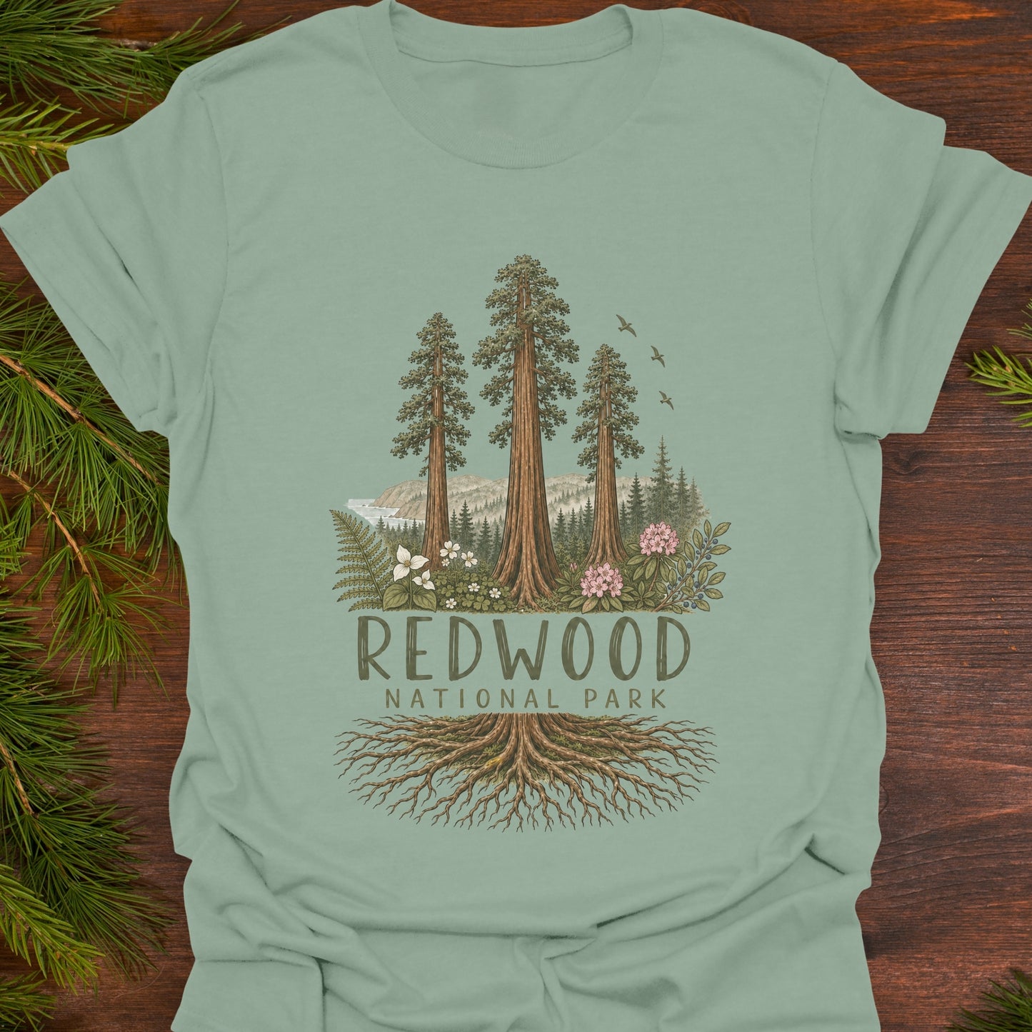 A sage green t-shirt laid flat on a dark wood background with pine branches in the corners. The shirt features a graphic of three tall redwood trees, mountains, birds, and the text 'REDWOOD NATIONAL PARK' with roots extending below.