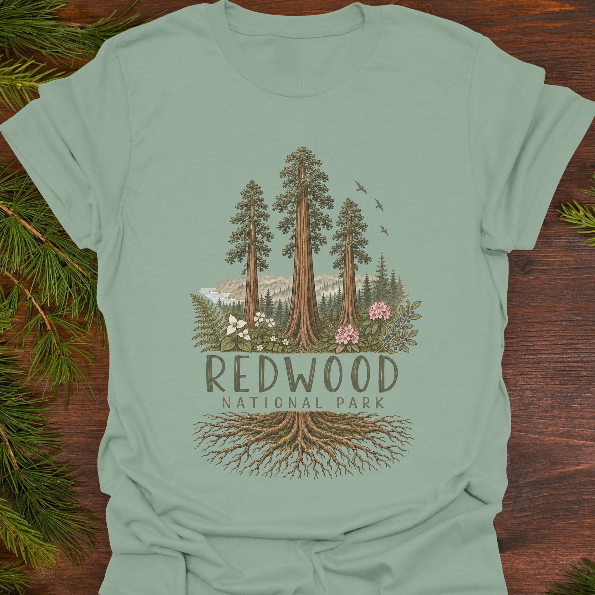 A sage green t-shirt laid flat on a dark wood background with pine branches in the corners. The shirt features a graphic of three tall redwood trees, mountains, birds, and the text 'REDWOOD NATIONAL PARK' with roots extending below.