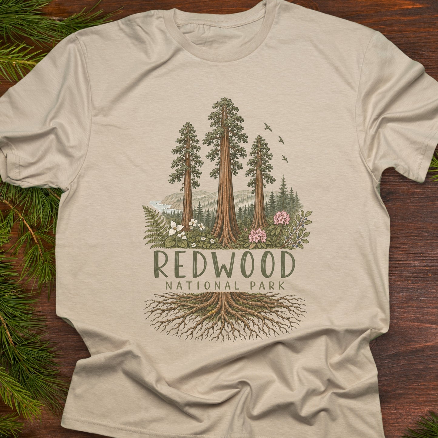 A beige crewneck t-shirt featuring a detailed graphic of Redwood National Park with redwood trees, forest foliage, and roots, laid flat on a dark wood background with pine branches.
