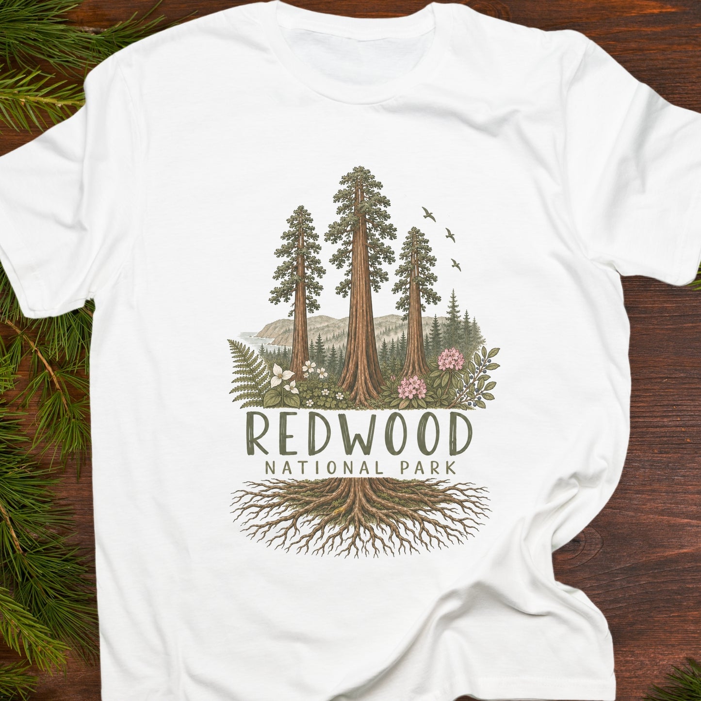 A white short-sleeve T-shirt laid flat on a dark wood surface with pine branches around the edges. The shirt features a large front graphic showing tall redwood trees, mountains, ferns, birds, and the text 'REDWOOD NATIONAL PARK' with tree roots illustrated below.