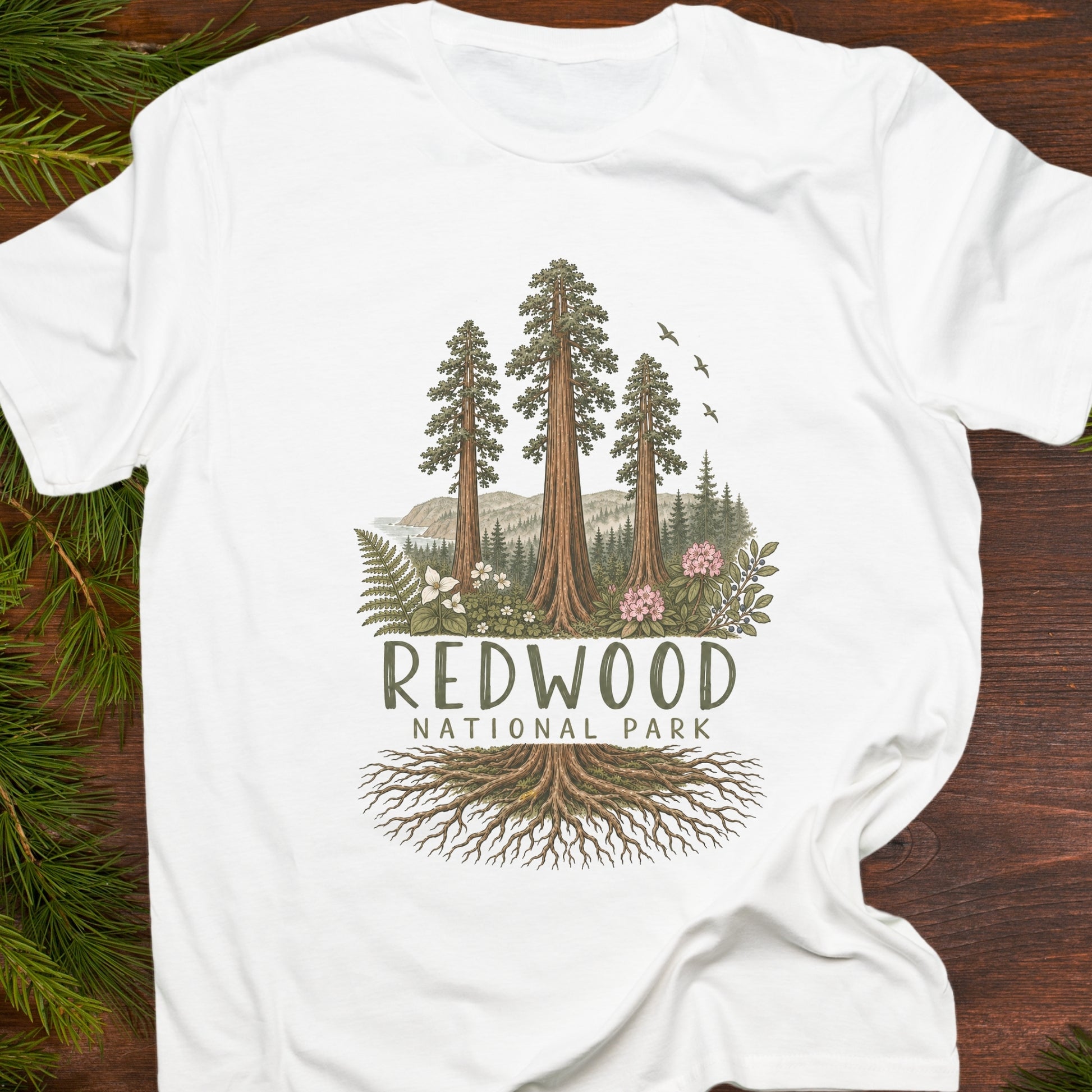 A white short-sleeve T-shirt laid flat on a dark wood surface with pine branches around the edges. The shirt features a large front graphic showing tall redwood trees, mountains, ferns, birds, and the text 'REDWOOD NATIONAL PARK' with tree roots illustrated below.