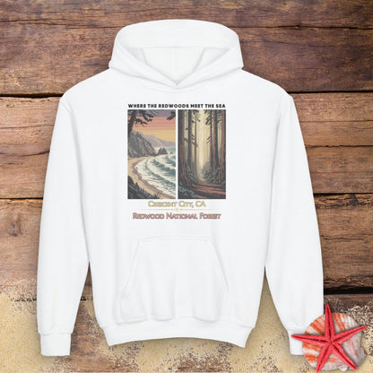 A white pullover hoodie laid flat on a rustic wooden background, featuring a vintage-style travel graphic of Redwood National Forest in Clear Creek City, CA. The graphic includes a coastal scene on the left and a forest scene on the right, with text reading 'WHERE THE REDWOODS MEET THE SEA' and 'REDWOOD NATIONAL FOREST'.