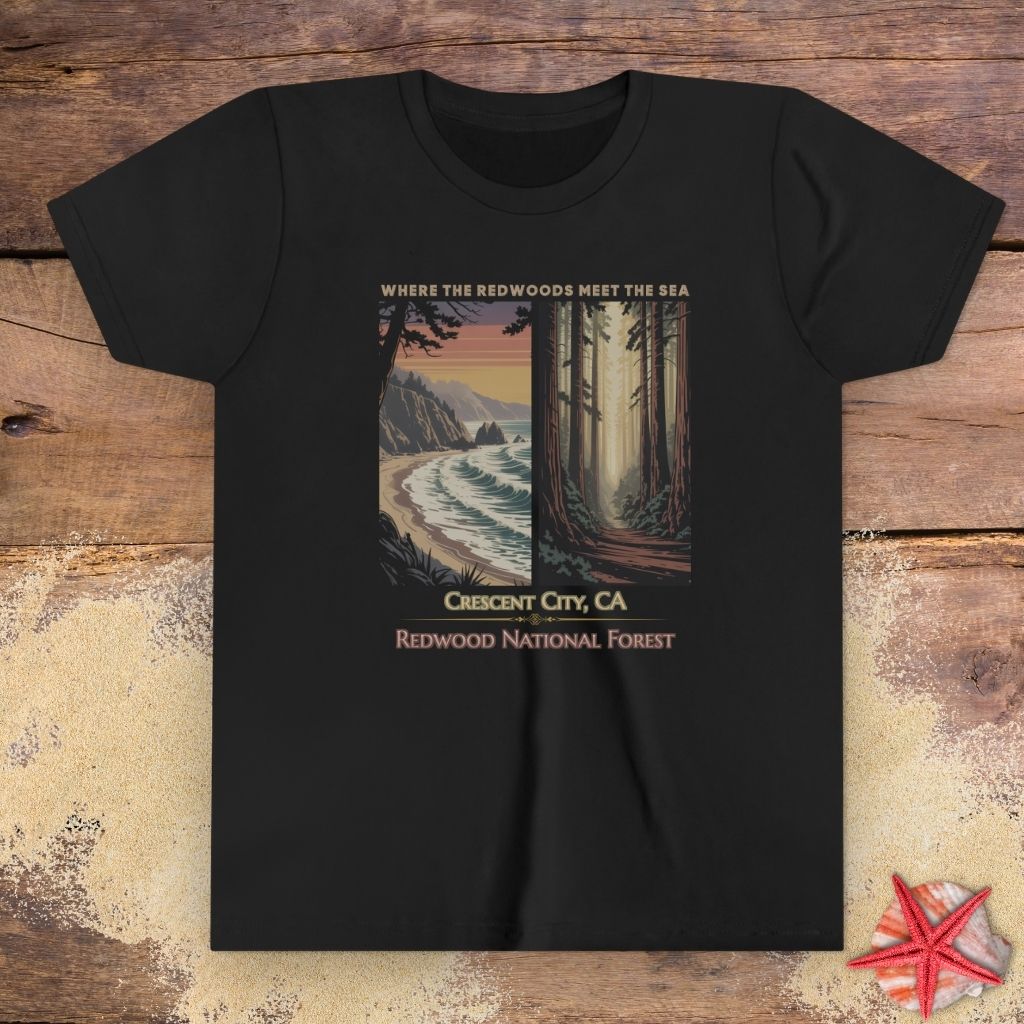 A black short-sleeve crewneck child t-shirt featuring a vintage-style travel poster graphic in the center depicting a coastal scene on the left and a redwood forest on the right, with the text 'WHERE THE OCEAN MEETS THE SEA', 'Crescent City, CA', and 'REDWOOD NATIONAL FOREST'.