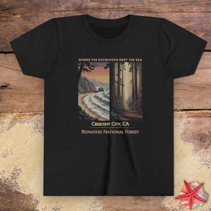 A black short-sleeve crewneck child t-shirt featuring a vintage-style travel poster graphic in the center depicting a coastal scene on the left and a redwood forest on the right, with the text 'WHERE THE OCEAN MEETS THE SEA', 'Crescent City, CA', and 'REDWOOD NATIONAL FOREST'.