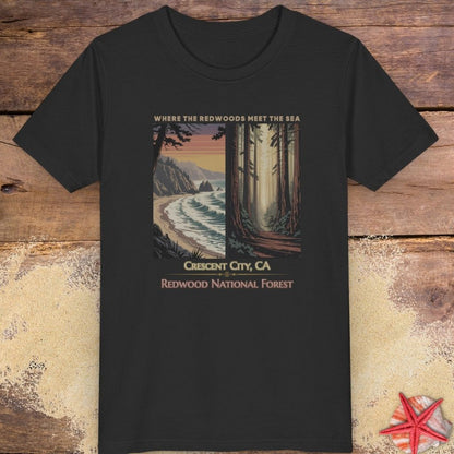 A black short-sleeve crewneck child t-shirt featuring a vintage-style graphic print of Crescent City, CA, showing a coastal scene and redwood forest with the text 'WHERE THE OCEAN MEETS THE SEA' and 'REDWOOD NATIONAL FOREST', displayed flat on a rustic wooden background.