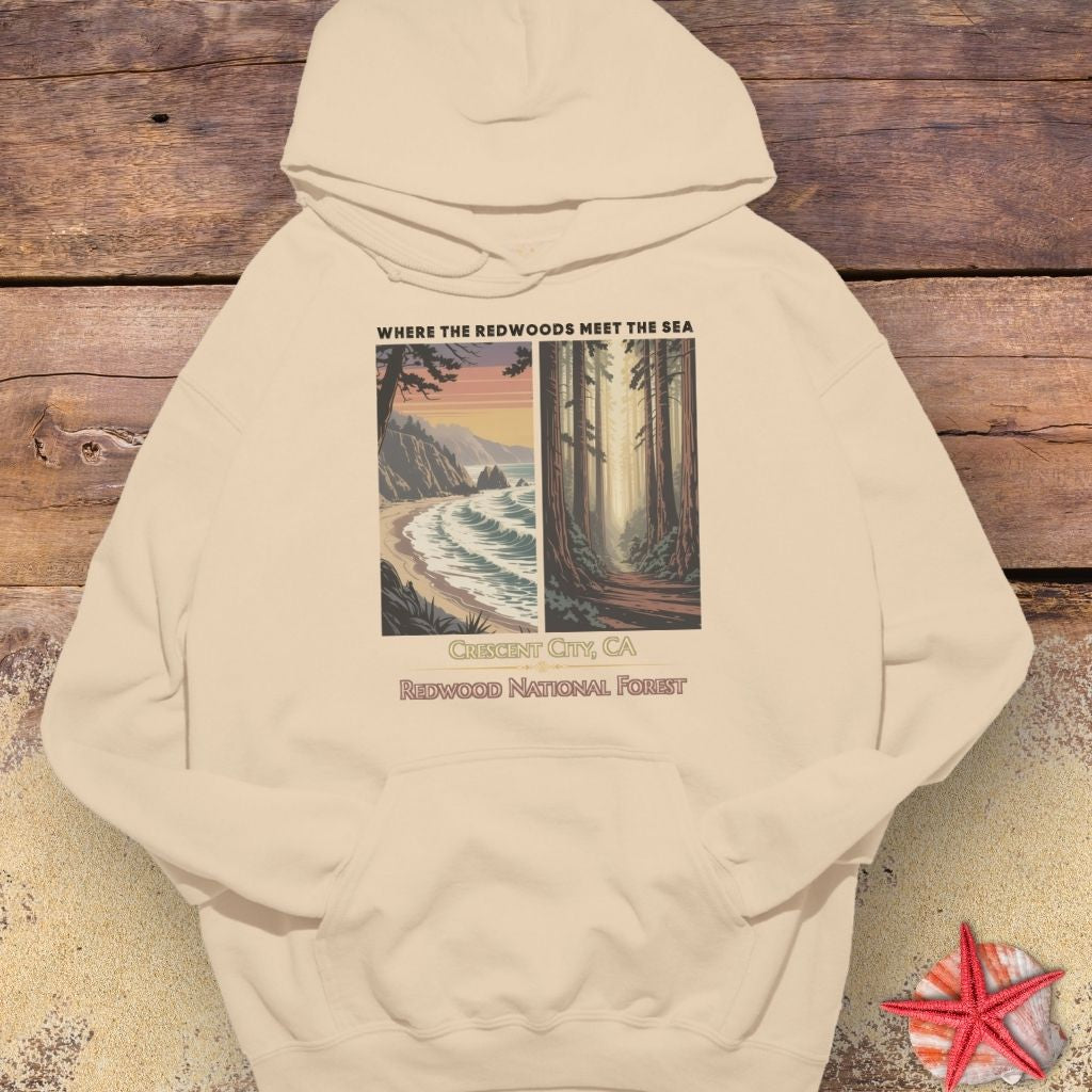 A beige pullover hoodie laid flat on a rustic wooden surface with a small red starfish prop at the bottom right. The hoodie features a large front graphic split into two scenic panels: a beach and ocean on the left, and a forest path on the right, with the text 'WHERE THE REDWOODS MEET THE SEA' above and 'CERRISSET CITY, CA / REDWOOD NATIONAL FOREST' below.