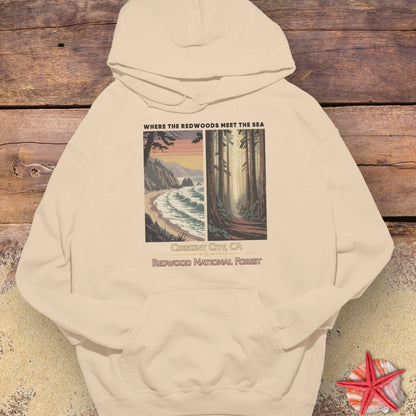 A beige pullover hoodie laid flat on a rustic wooden surface with a small red starfish prop at the bottom right. The hoodie features a large front graphic split into two scenic panels: a beach and ocean on the left, and a forest path on the right, with the text 'WHERE THE REDWOODS MEET THE SEA' above and 'CERRISSET CITY, CA / REDWOOD NATIONAL FOREST' below.
