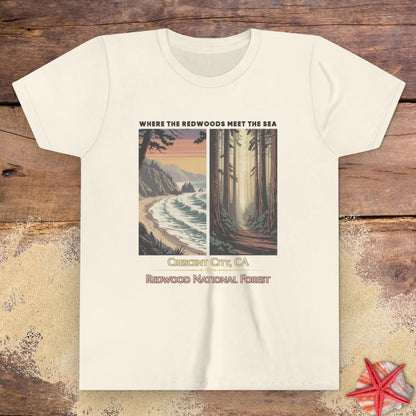 A beige short-sleeve crewneck child t-shirt featuring a graphic print of a coastal landscape and a forest, with the text 'WHERE THE REDWOODS MEET THE SEA', 'CINCUT CITY, CA', and 'REDWOOD NATIONAL FOREST'.