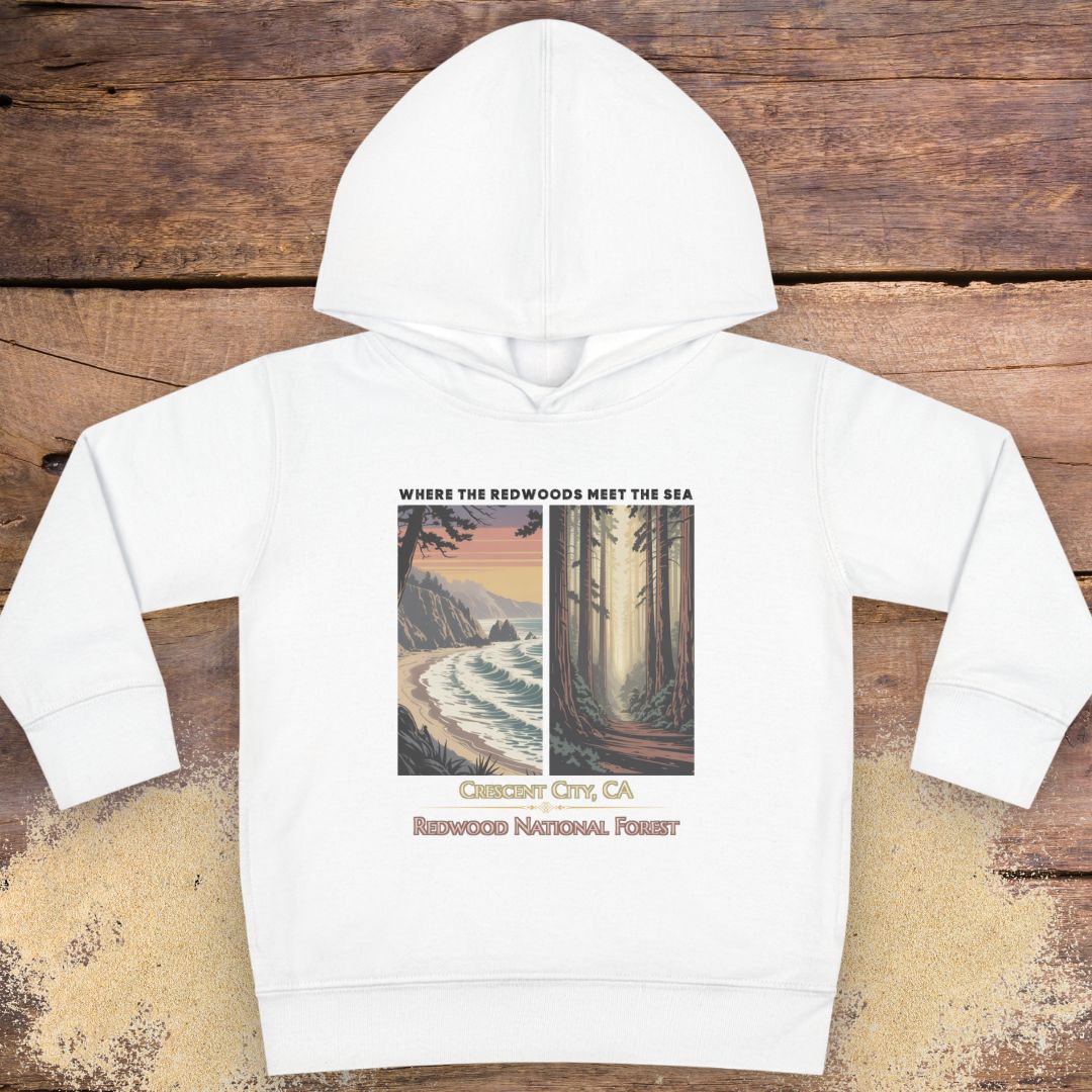 A white kids' pullover hoodie with a graphic print on the front, laid flat on a rustic wooden background. The graphic features a coastal scene on the left and a redwood forest on the right, with text reading 'WHERE THE REDWOODS MEET THE SEA', 'Carmen, CA', and 'REDWOOD NATIONAL FOREST'.