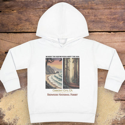 A white kids' pullover hoodie with a graphic print on the front, laid flat on a rustic wooden background. The graphic features a coastal scene on the left and a redwood forest on the right, with text reading 'WHERE THE REDWOODS MEET THE SEA', 'Carmen, CA', and 'REDWOOD NATIONAL FOREST'.
