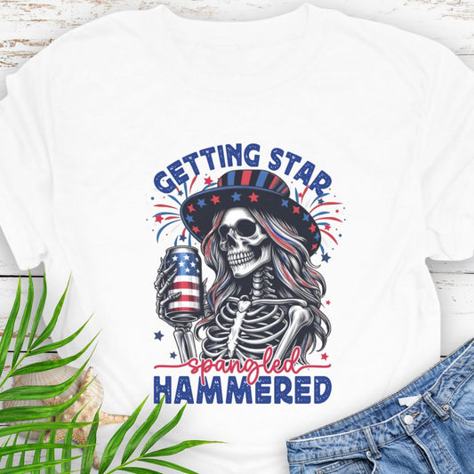 White T-shirt graphic of a woman skeleton wearing a stars-and-stripes hat, holding an American flag beer can with the phrase “Getting Star Spangled Hammered.