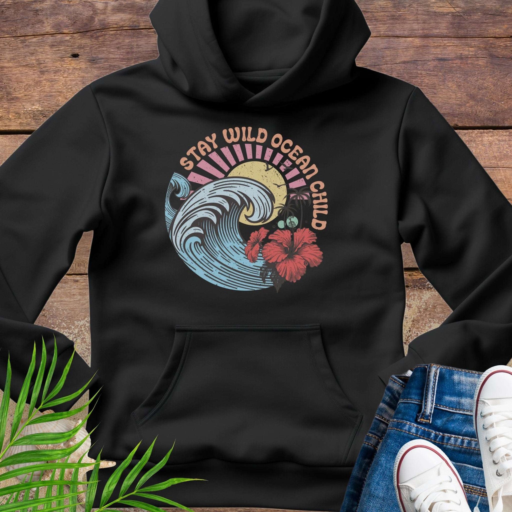 Black hoodie with retro beach artwork featuring a stylized wave, palm trees, hibiscus flowers, and the phrase "Stay Wild Ocean Child" in sunset colors.