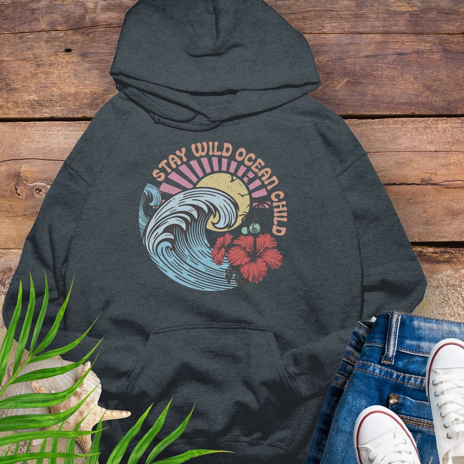 Dark gray hoodie with retro beach artwork featuring a stylized wave, palm trees, hibiscus flowers, and the phrase "Stay Wild Ocean Child" in sunset colors.