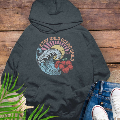 Dark gray hoodie with retro beach artwork featuring a stylized wave, palm trees, hibiscus flowers, and the phrase "Stay Wild Ocean Child" in sunset colors.