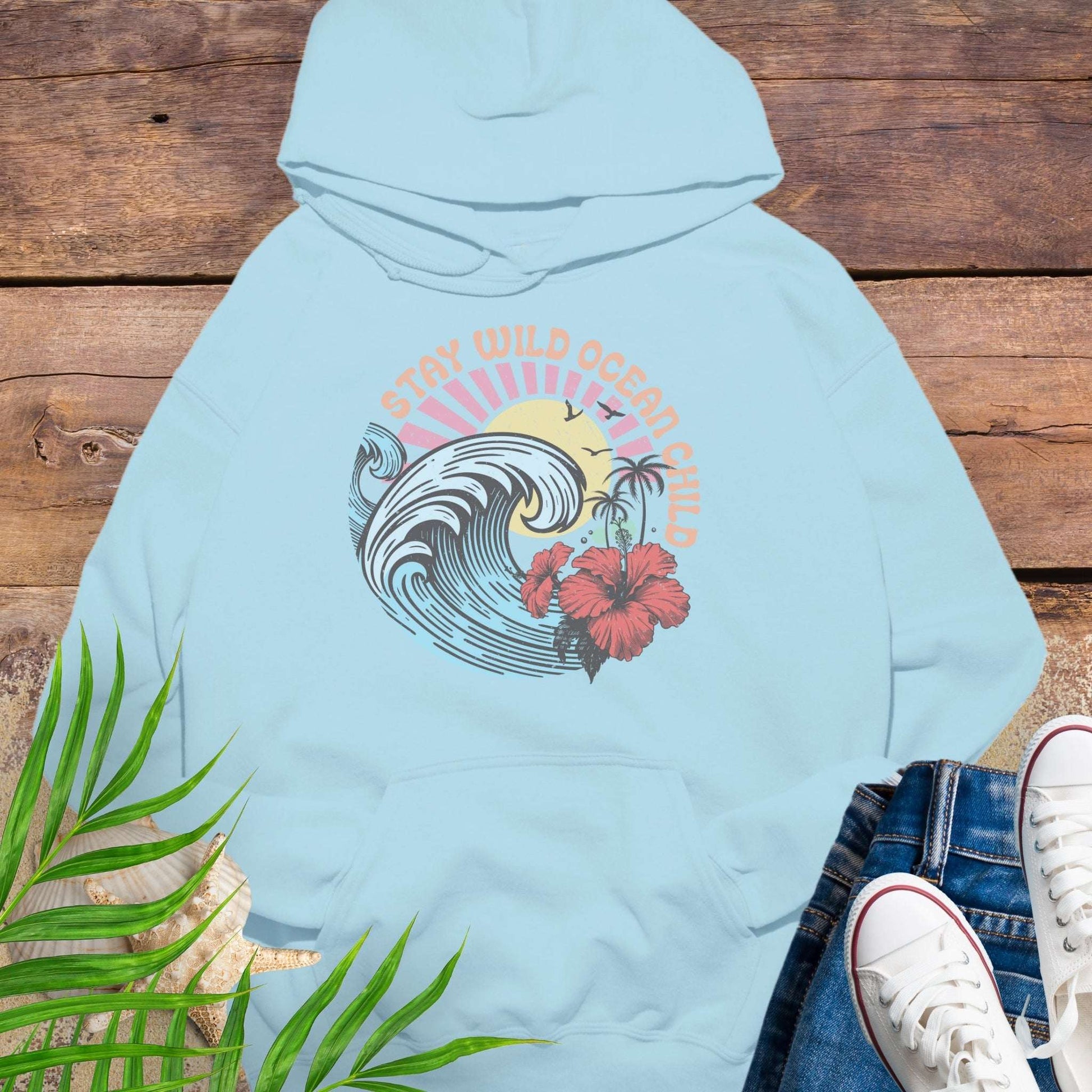 Light blue hoodie with retro beach artwork featuring a stylized wave, palm trees, hibiscus flowers, and the phrase "Stay Wild Ocean Child" in sunset colors.