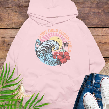 Pink hoodie with retro beach artwork featuring a stylized wave, palm trees, hibiscus flowers, and the phrase "Stay Wild Ocean Child" in sunset colors.