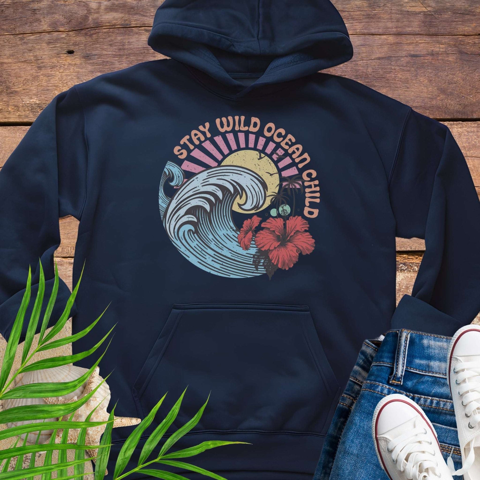Navy blue hoodie with retro beach artwork featuring a stylized wave, palm trees, hibiscus flowers, and the phrase "Stay Wild Ocean Child" in sunset colors.