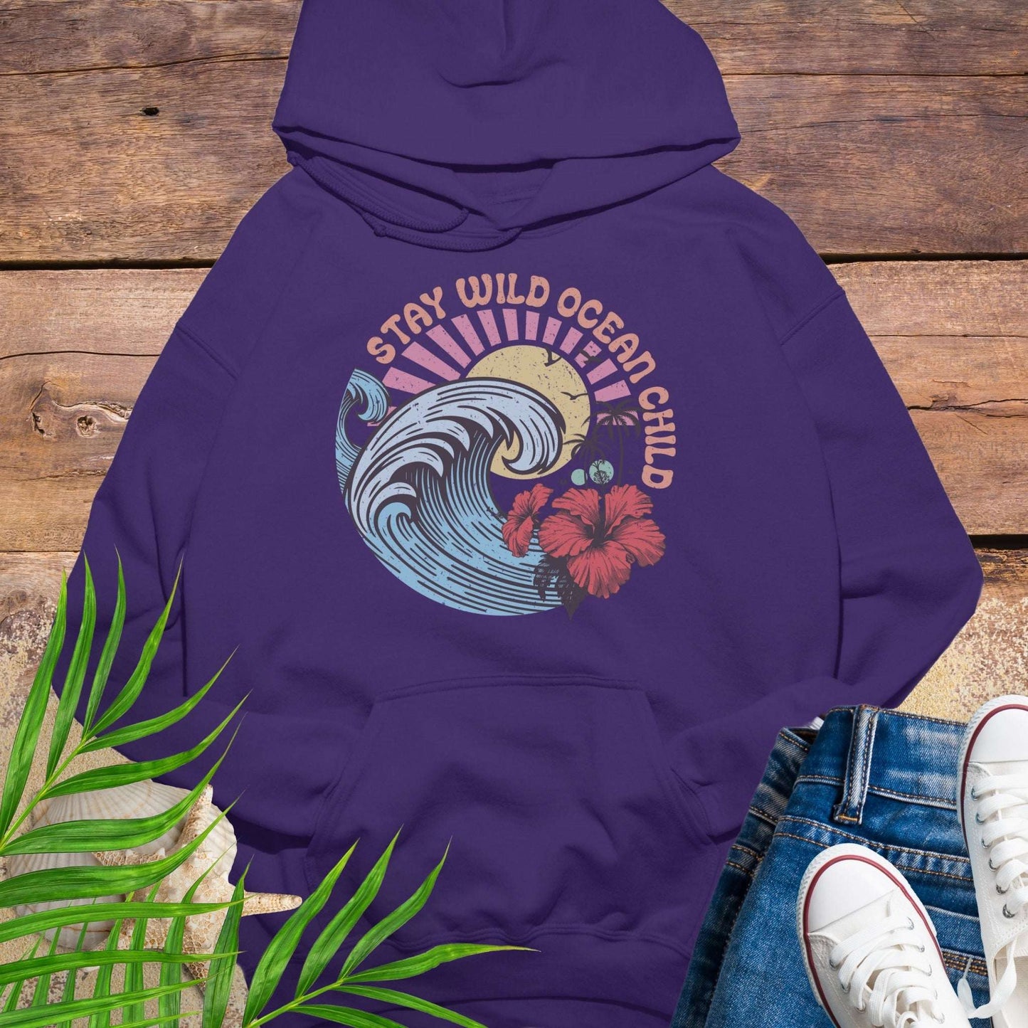 Purple hoodie with retro beach artwork featuring a stylized wave, palm trees, hibiscus flowers, and the phrase "Stay Wild Ocean Child" in sunset colors.