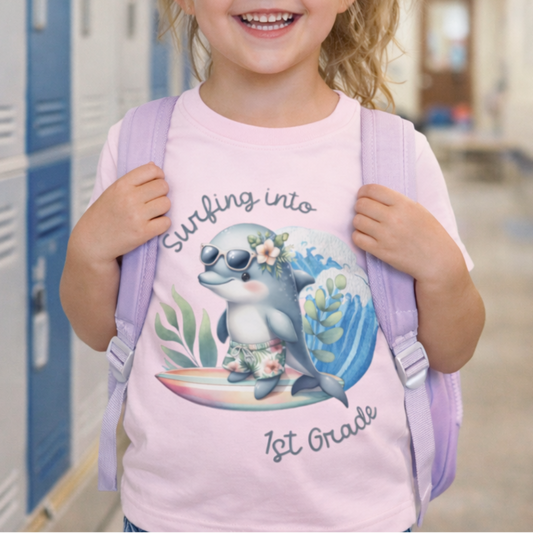 Young girl in a school hallway wearing a pink shirt with a dolphin graphic with text Surfing into 1st Grade.