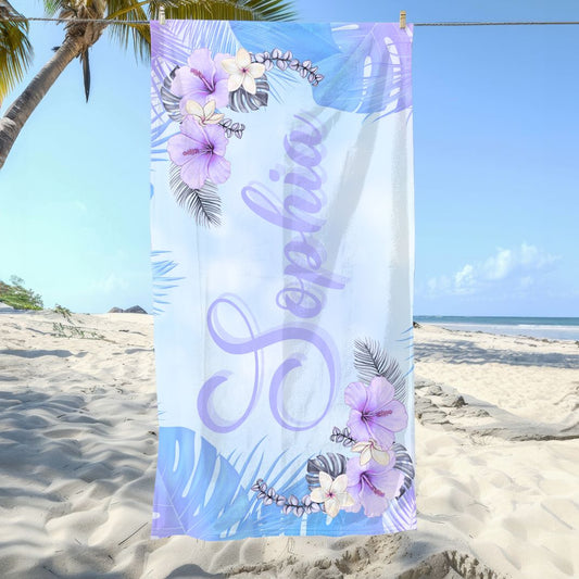 Beach towel with soft blue and purple background, hibiscus flowers and greenery in corners, and personalized name in script font.