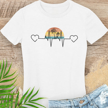White t-shirt with a heart rate design featuring a sunset and palm trees on a wooden surface with green leaves.