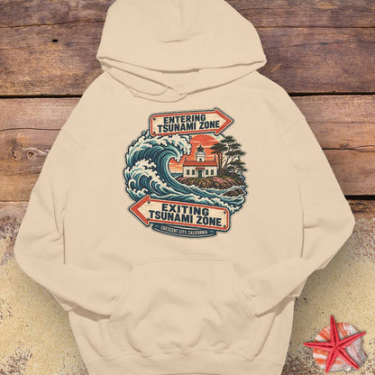 A beige Crescent City California hoodie design featuring a crashing ocean wave, coastal lighthouse, and vintage entering and exiting tsunami zone signs.