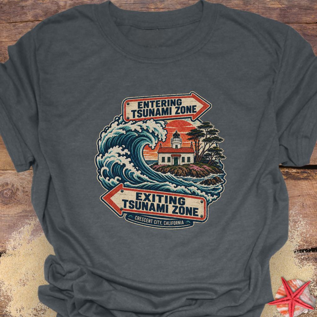 A dark gray heather t-shirt featuring a vintage-style graphic of a tsunami wave and lighthouse with retro signs reading 'ENTERING TSUNAMI ZONE' and 'EXITING TSUNAMI ZONE'.